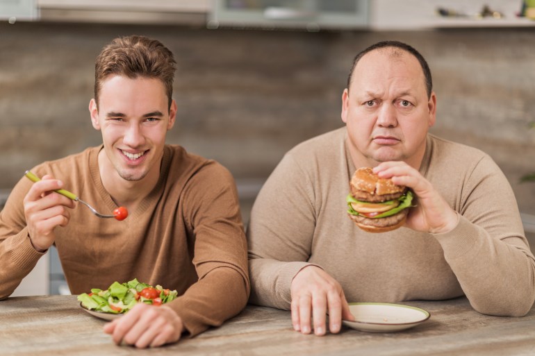 أكل طعام صحي و غير صحي ، الوجبات السريعة ، وجبات سريعة ، مطاعم أمريكية healthy food versus unhealthy food Two men eating together in the kitchen and looking at camera. Fat sad man is eating a hamburger and the other one is smiling while eating salad.