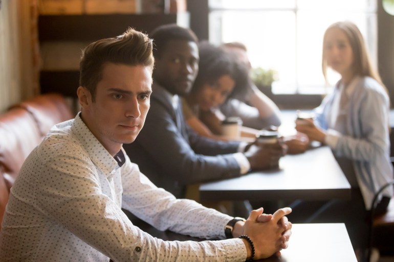 Diverse people looking at thoughtful frustrated man in cafe, sad misunderstood guy feels offended ignoring multiracial friends sitting alone at table, excluded outstand person suffers from bullying