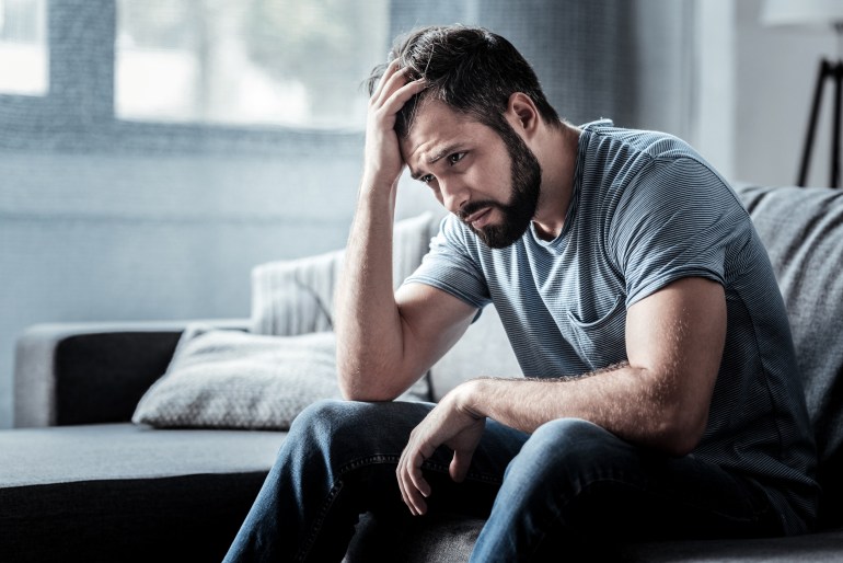 Unpleasant pain. Sad unhappy handsome man sitting on the sofa and holding his forehead while having headache