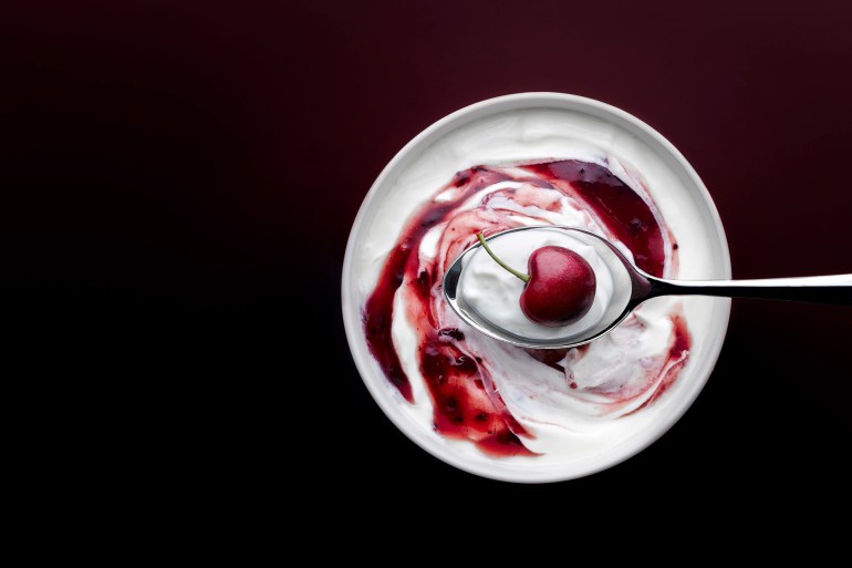 CLOSE UP SHOT OF YOGURT AND CHERRY ON SPOON ABOVE A BOWL