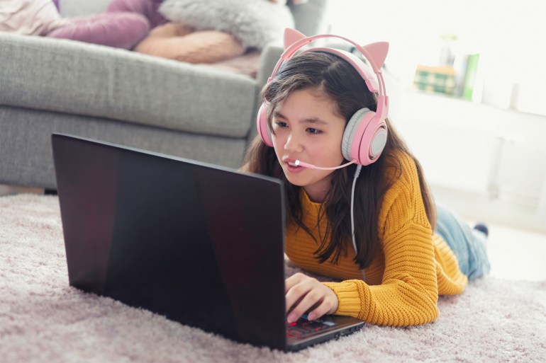 A girl laying down on a living room carpet and playing an online video game on a laptop computer while communicating using a headset with microphone.