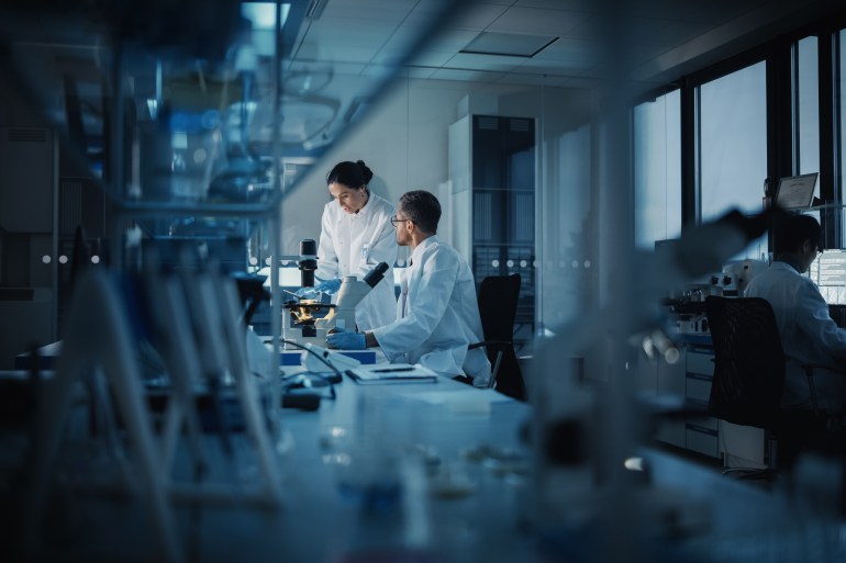 Modern Medical Research Laboratory: Two Scientists Working, Using Digital Tablet, Analyzing Test, Talking. Advanced Scientific Pharmaceutical Lab for Medicine, Biotechnology Development. Evening Time