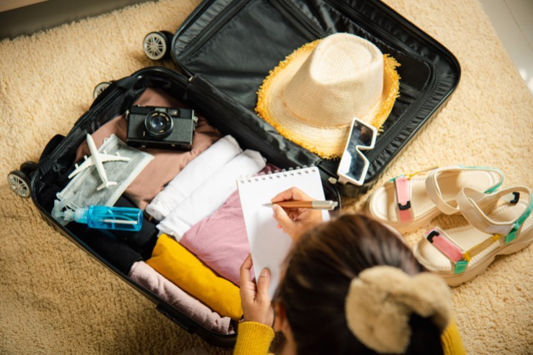 Making check list of things to pack for travel. Woman writing paper take note and packing suitcase to vacation writing paper list sitting on room, prepare clothes into luggage, Travel vacation travel