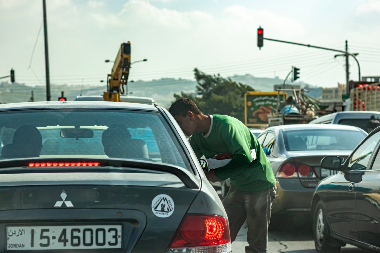 Amman, Jordan 03-29-2010: A poor young refugee boy is begging at the traffic light on a crowded road in Amman. The young Arabic man taps on window of cars stopping at red light and asks for money .