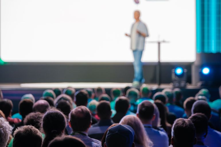 Speaker delivering a presentation to a large audience at a business conference in a modern venue. Engaged attendees listen attentively.