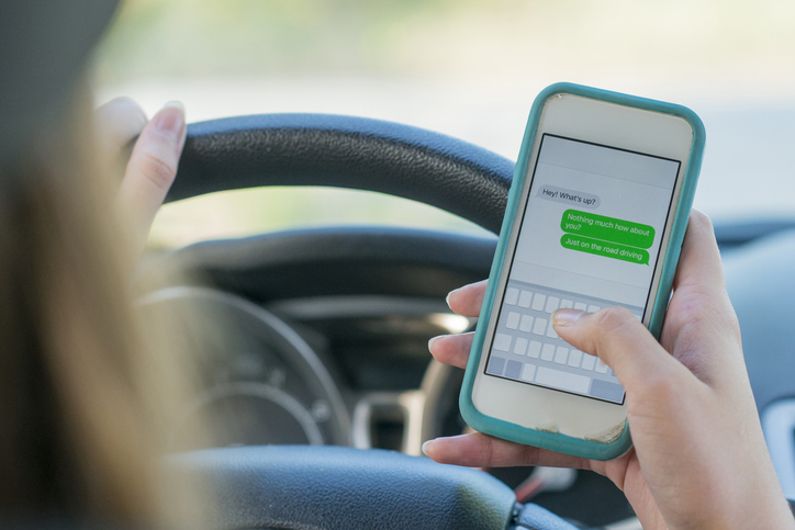 A Caucasian teenage girl is sitting in the driver's seat of her new black car. She is sending a text message to her friend while driving. She is driving recklessly.