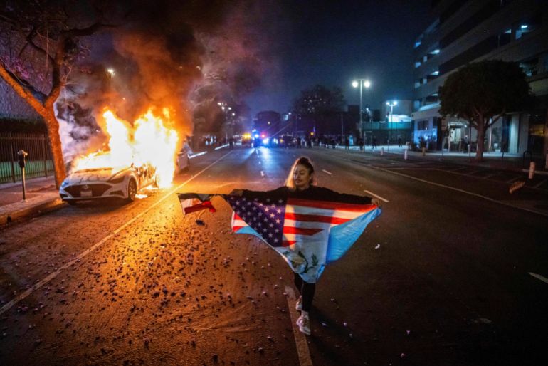 A person carrying multiple flags walks past a burning car during protests over the Trump administration's immigration raids in Los Angeles, Monday, June 9, 2025. (AP Photo/Ethan Swope)