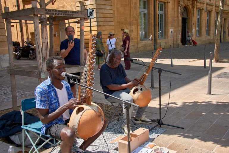 Aix, Provence, France; May 31, 2025, two Black musicians sitting, focused on playing the kora, a traditional African stringed instrument, on a pedestrian street in Aix-en-Provence, France. Their instruments, made of calabash and leather, and their microphones are in the foreground, while in the background, a lively urban environment with people strolling and trees providing shade can be seen. The warm sunlight highlights the musicians' skin tones and the materials of the instruments, conveying an atmosphere of authenticity, talent, and cultural richness, ideal for projects about music, African culture, street art, tourism, or cultural diversity.