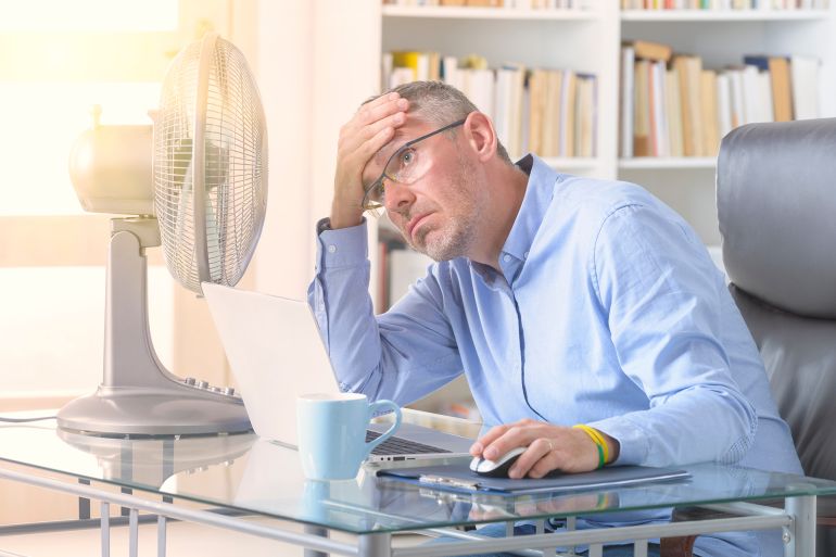 Man suffers from heat while working in the office and tries to cool off by the fan; Shutterstock ID 1775051300; purchase_order: aj; job: ; client: ; other:
