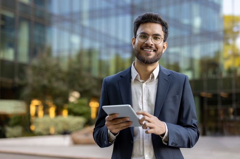Young businessman in suit using tablet outside modern glass office building. Confident professional smiling working on digital device. Concept of business, technology, success, and entrepreneurship.; Shutterstock ID 2482343185; purchase_order: aljazeera ; job: ; client: ; other: