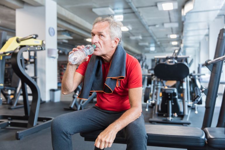 Mature man sitting at the gym and drinking water from bottle. Senior man taking a break after workout at rehab gym.
